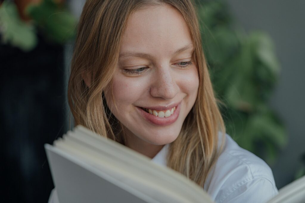 A woman with blonde hair and freckles smiles while reading a book indoors.